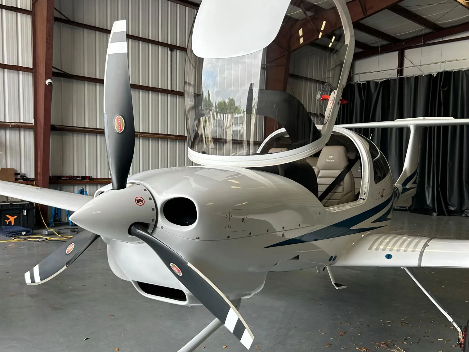 Front-side view of a white Diamond DA40 aircraft with blue stripes and its canopy open, parked in a hangar.