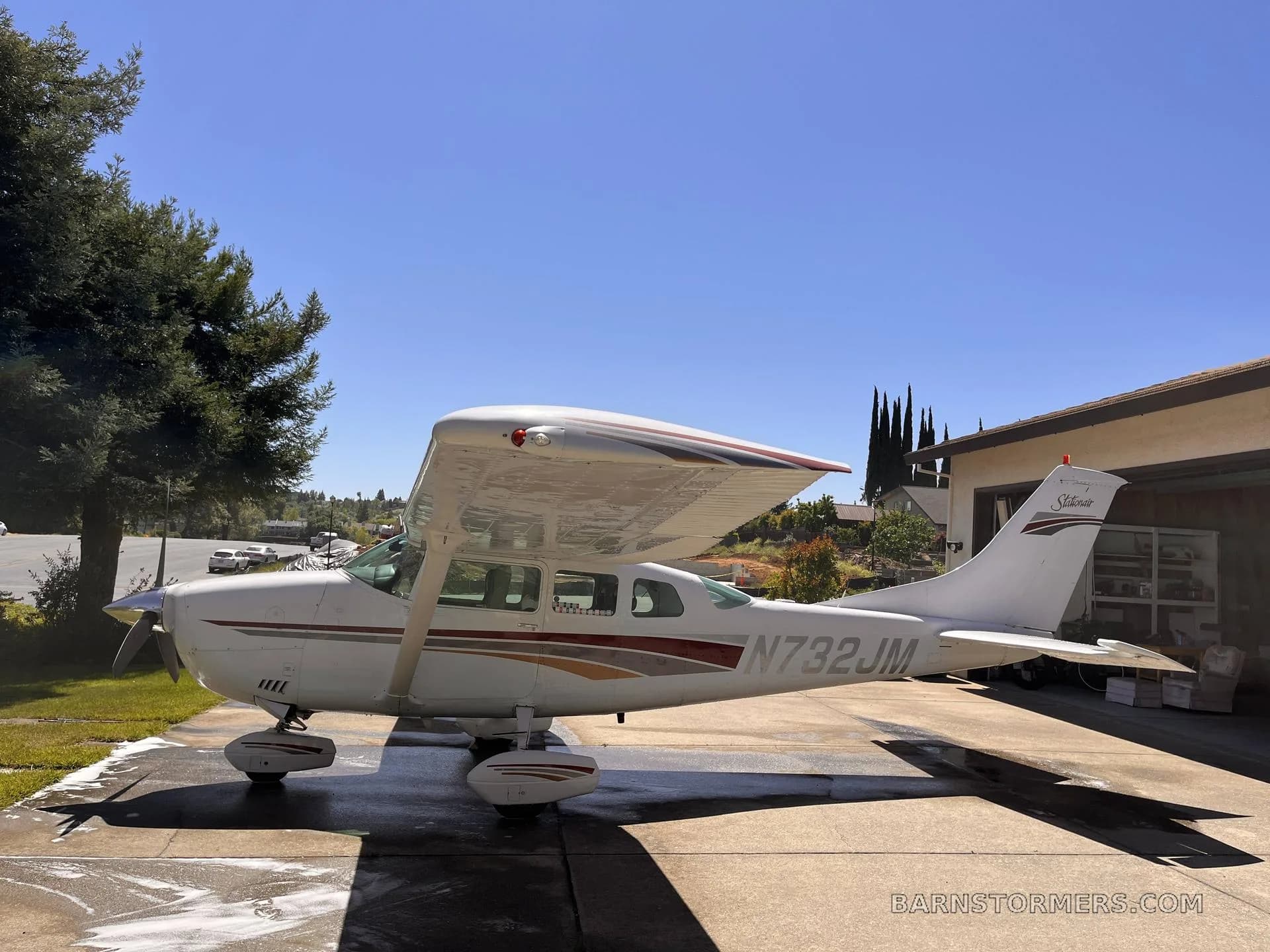 A white Cessna Skylane (N732JM) with red and gold stripes is parked on a wet driveway, viewed from the front left, under a clear blue sky.