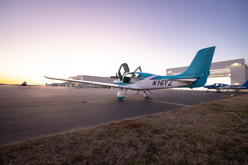 A turquoise and white Cirrus SR22 aircraft with its doors open, parked on an airfield next to hangars at sunset.