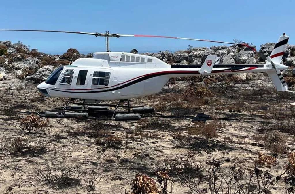 A white helicopter with black and red stripes parked in a rocky, dry landscape under a blue sky.