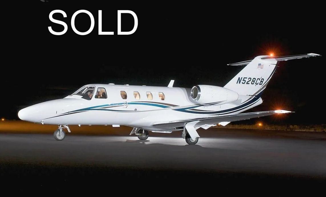 A white Cessna Citation business jet with blue and brown stripes is parked on a tarmac at night, with its navigation lights illuminated.