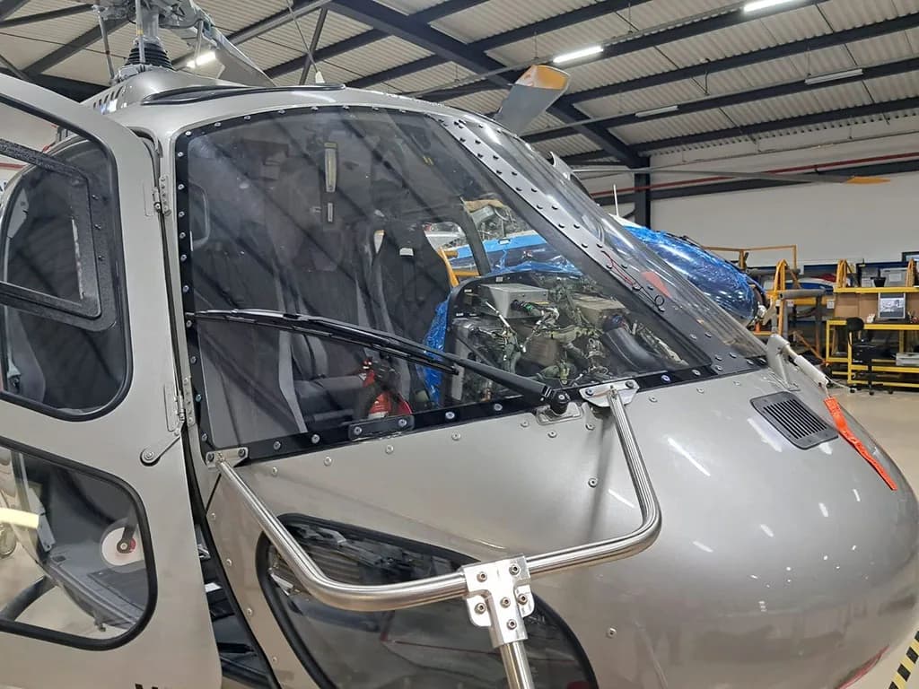 A close-up view of the front windshield and nose of a silver Airbus H125 helicopter inside a hangar.