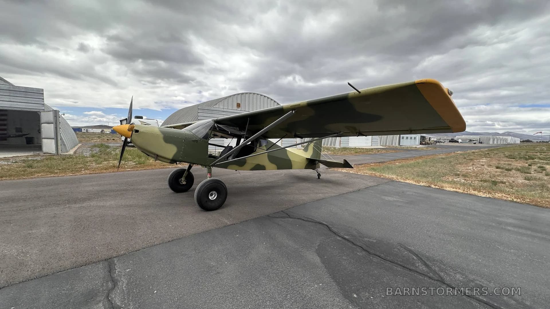 A light aircraft with camouflage paint and a yellow propeller spinner is parked on an asphalt runway with hangars in the background.