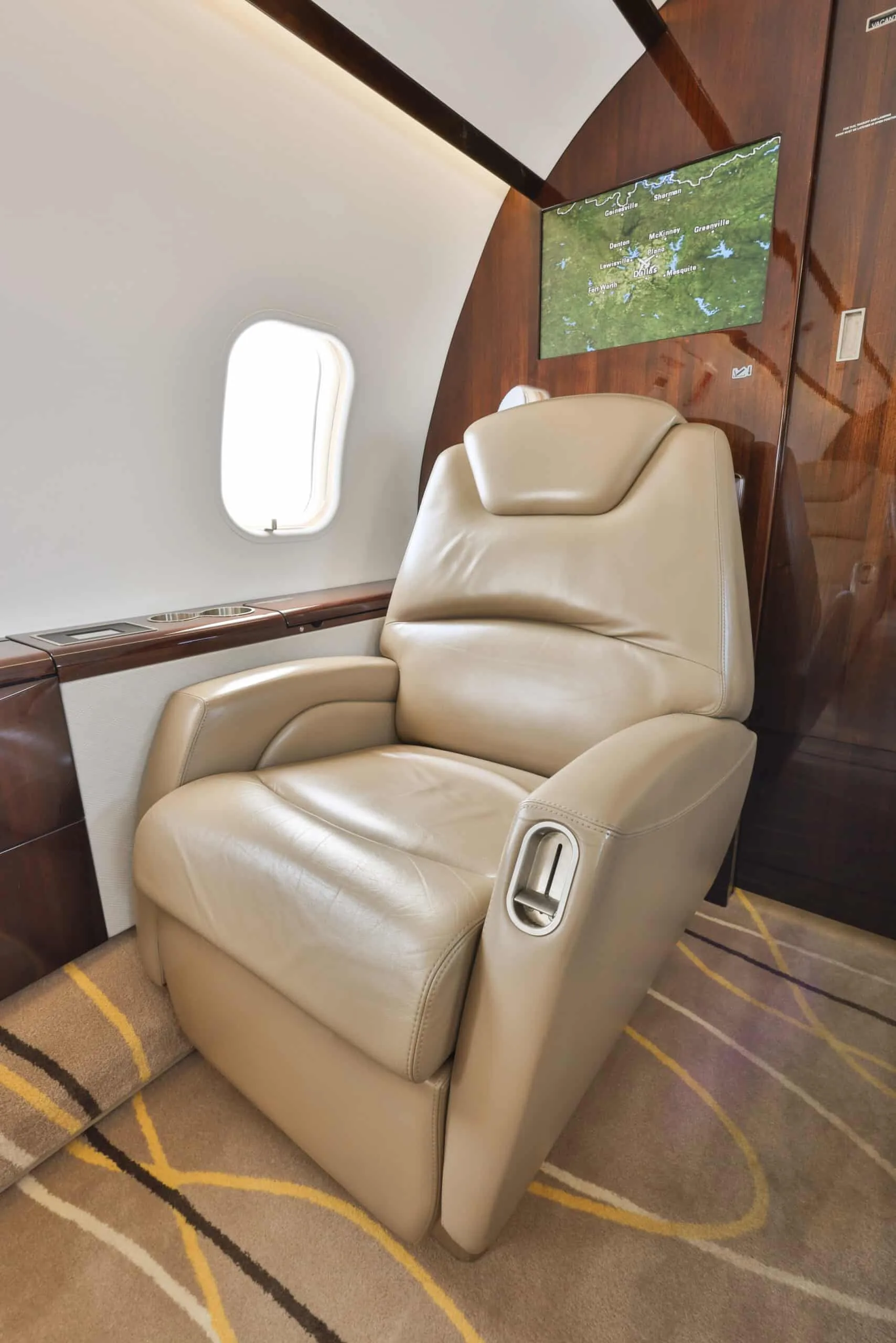 A single beige leather club seat is shown in the private jet cabin next to a window and a display screen showing a map.