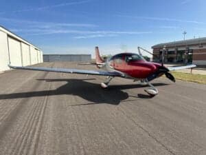 A Cirrus SR22T aircraft with a red and silver paint scheme is parked on a tarmac near a building.