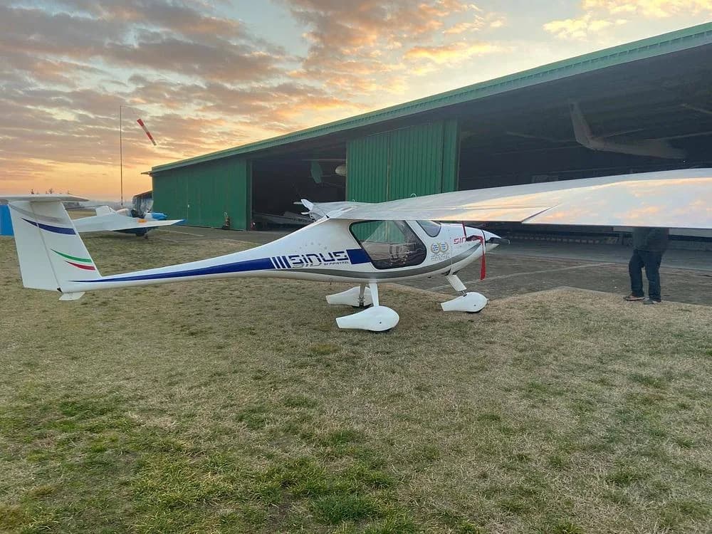 A white Pipistrel Sinus aircraft with blue and green tail stripes, parked on a grassy area in front of a green hangar at sunset.