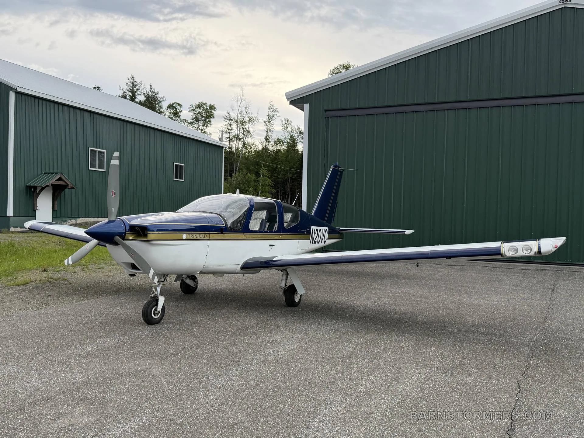 A blue and white Socata Trinidad TB20 aircraft, registration N20VC, parked on an asphalt ramp in front of green hangars.