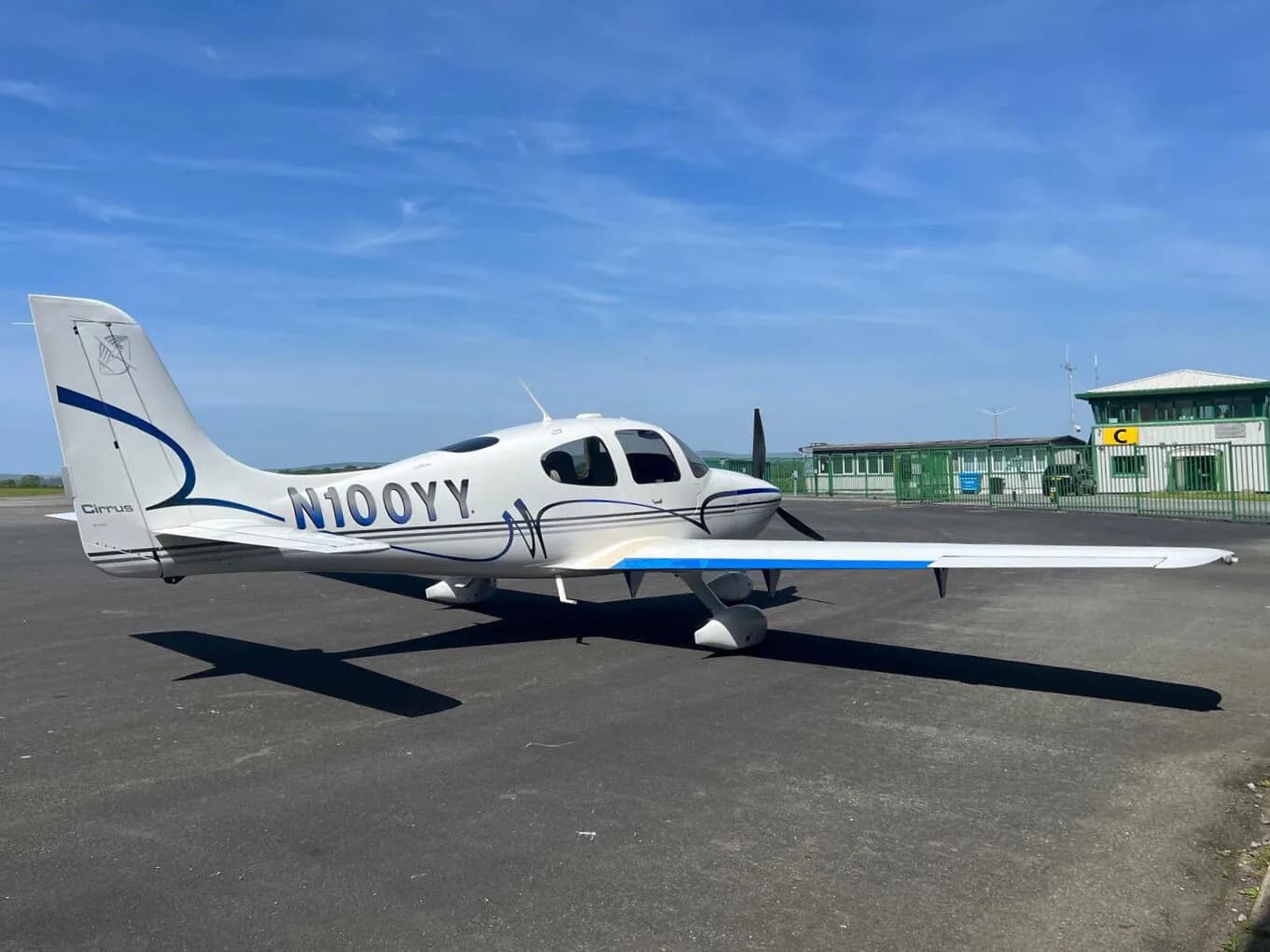 A white Cirrus SR22 aircraft with blue and grey stripes parked on an airfield tarmac under a blue sky.