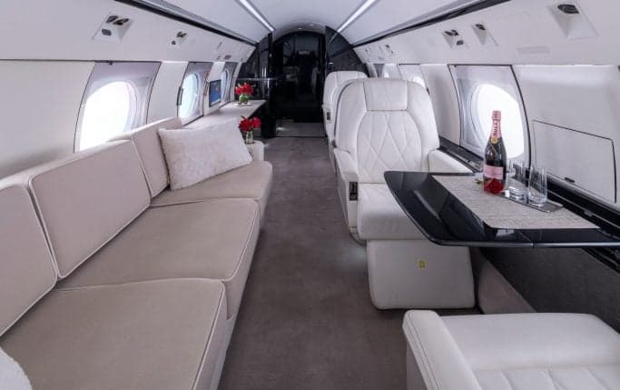 A view down the aisle of the aircraft cabin, featuring white leather seats and tables.