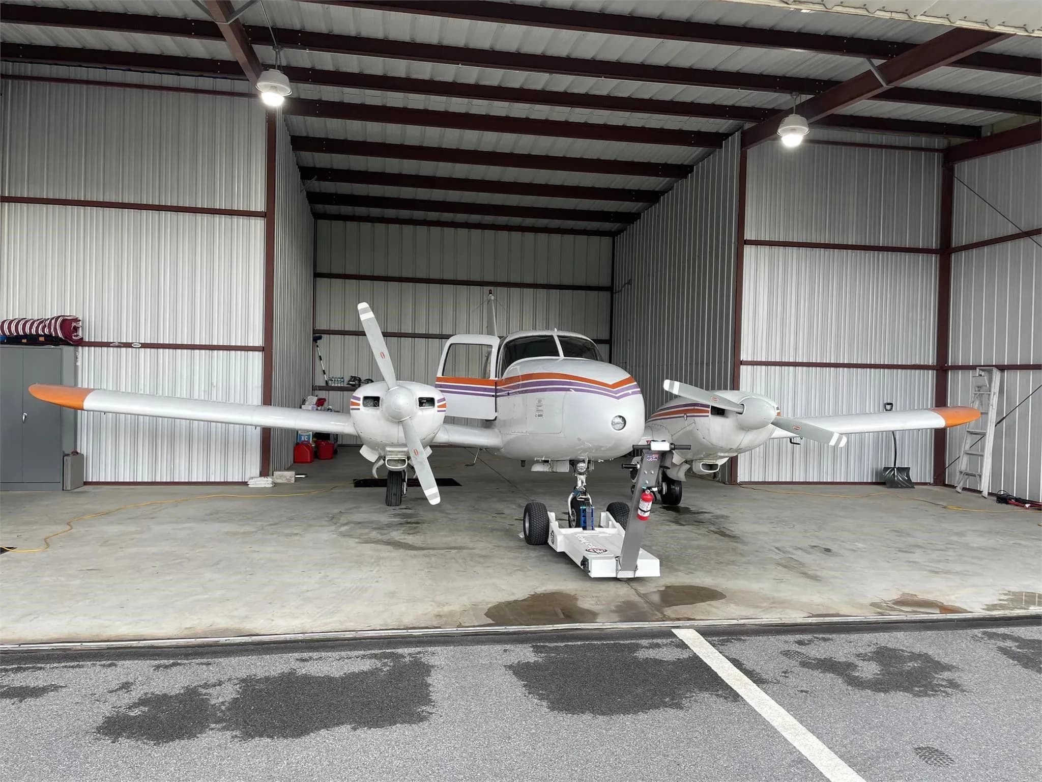 A full front view of the white Piper Aztec twin-engine aircraft with orange and purple stripes parked in a hangar.