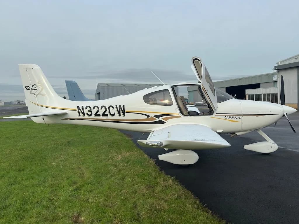A side view of a white Cirrus SR22 aircraft with N322CW registration and open gull-wing doors, parked on a tarmac.