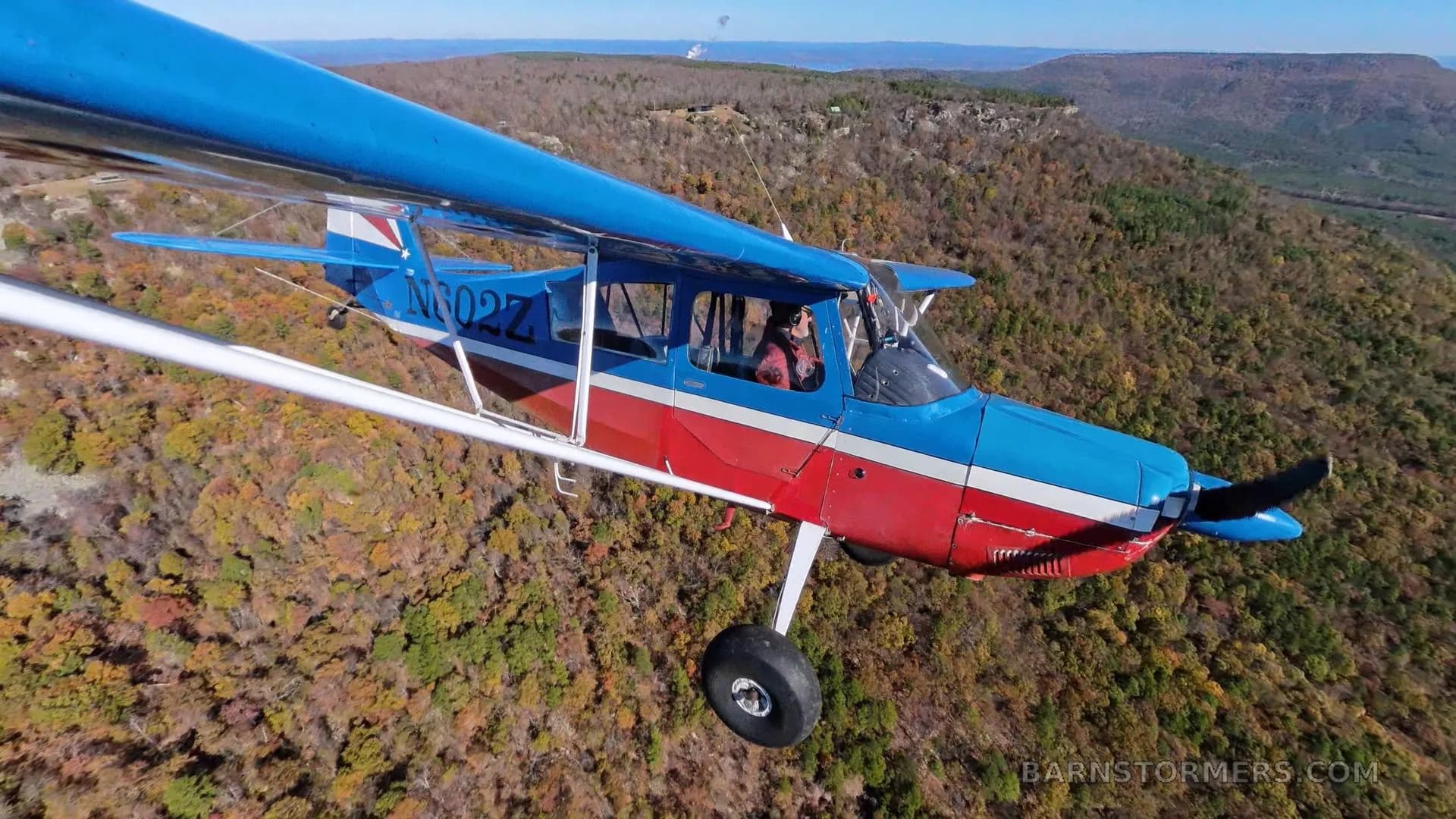 A red and blue high-wing aircraft with registration N602Z flies over a forested mountain landscape.