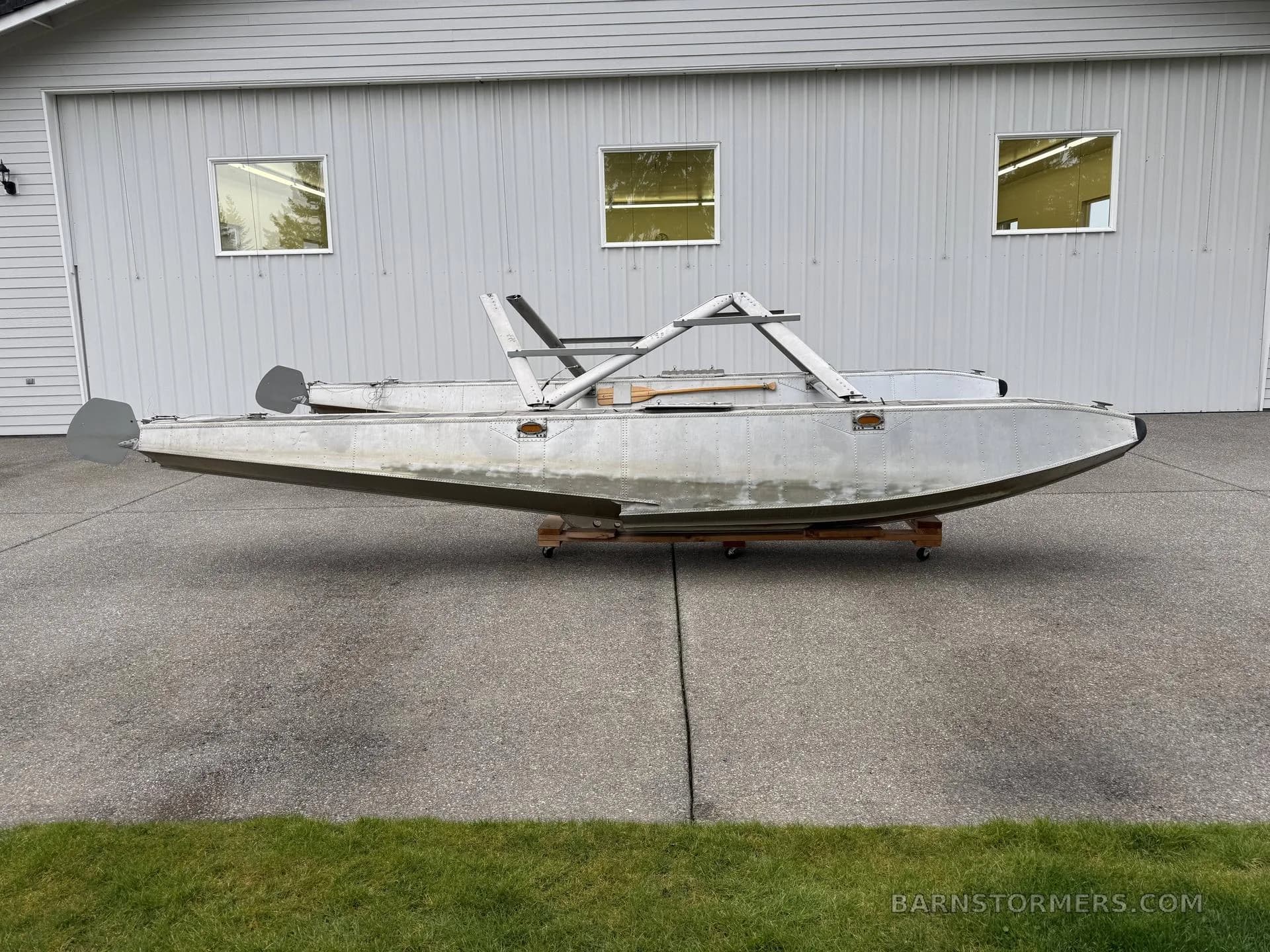 A pair of aluminum aircraft floats with mounting struts, seen from the side, on a concrete driveway in front of a white building.