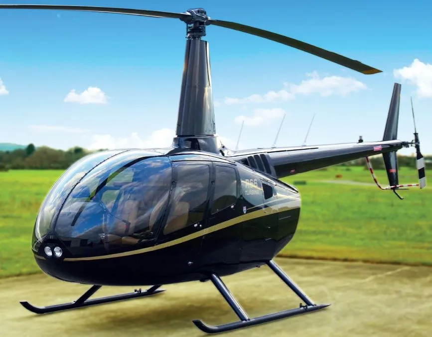 A black Robinson R66 Turbine helicopter with a gold stripe is parked on a grassy field under a blue sky.