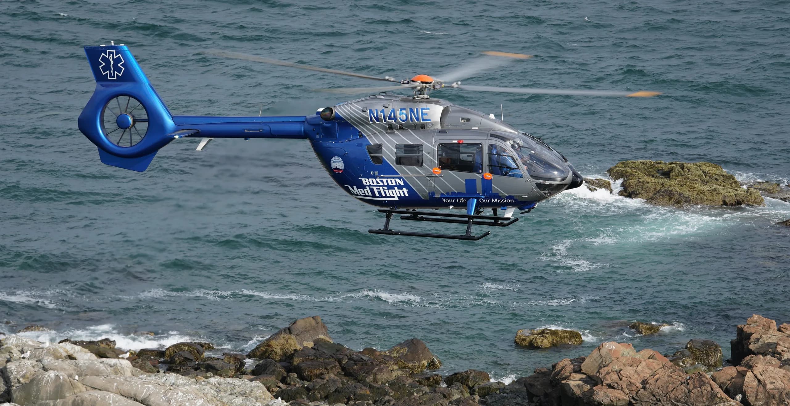 A blue and gray Boston MedFlight helicopter flying over the ocean.