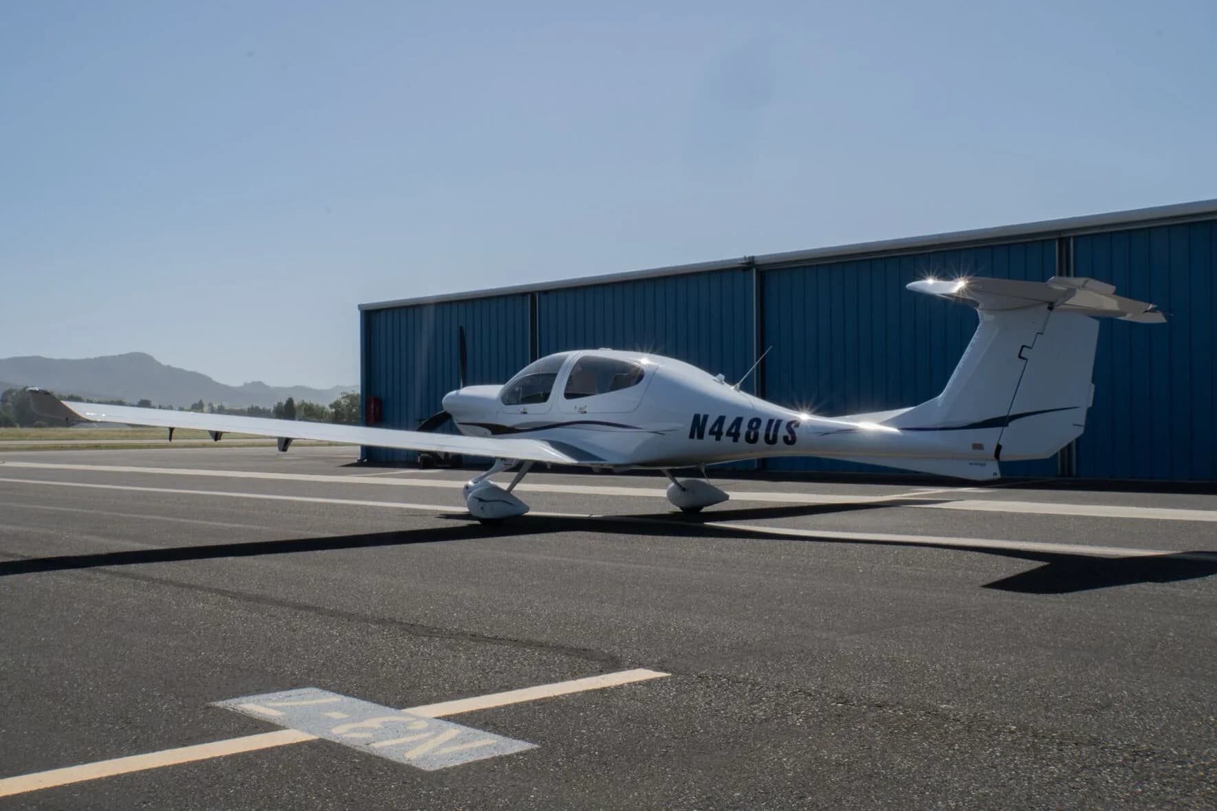 A white Diamond DA40 Star XL aircraft with purple and grey stripes, registration N448US, parked on an tarmac in front of a blue hangar.