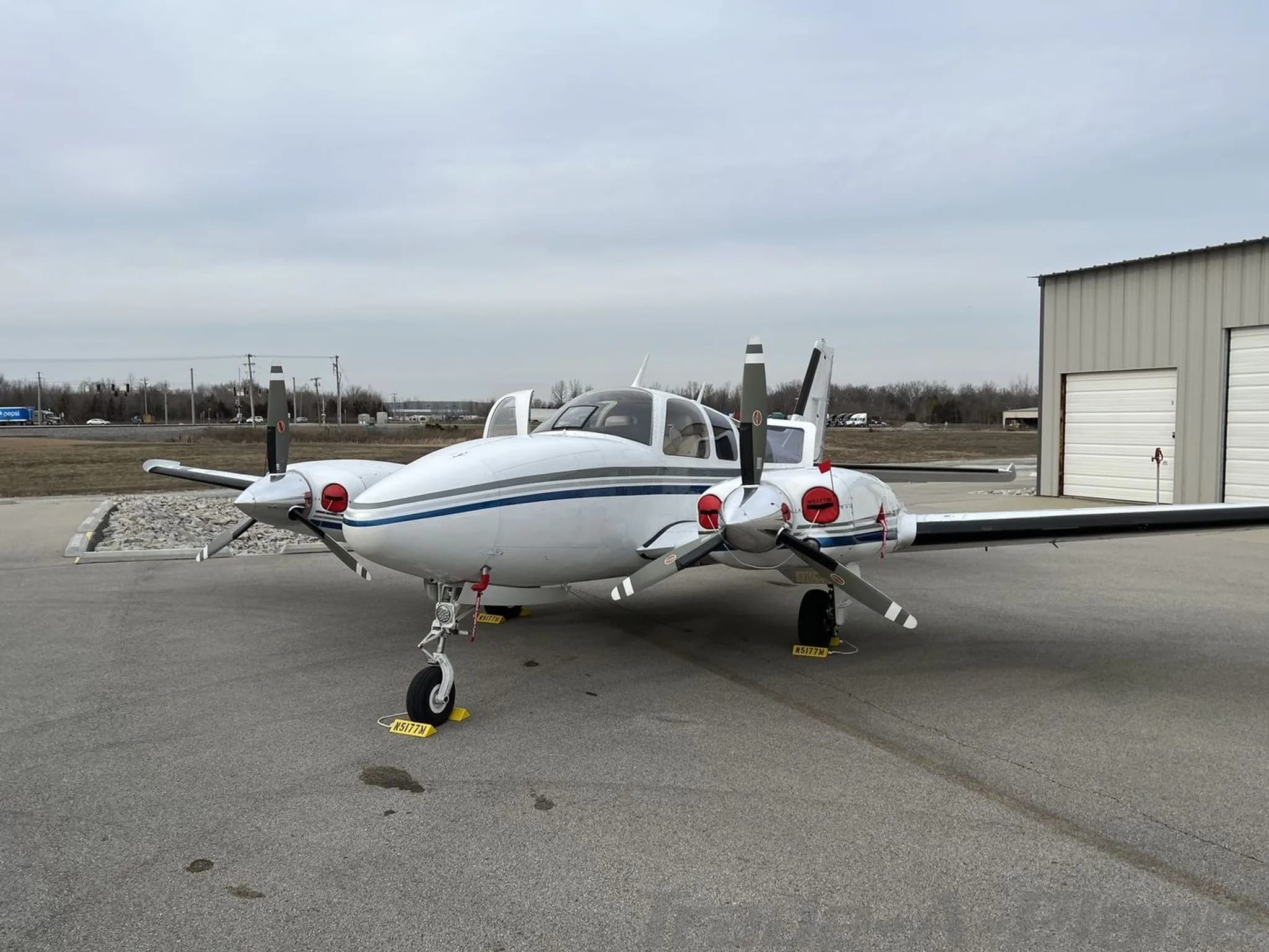 A head-on view of the white and blue twin-engine Beechcraft Baron N5177M with its main cabin door open, parked on an airfield.