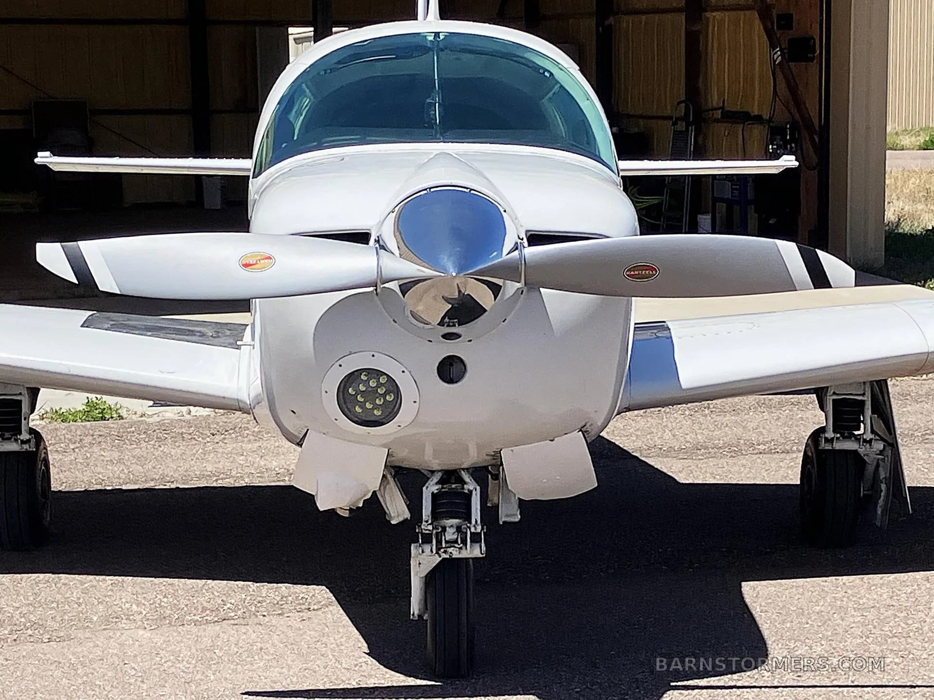 A head-on view of a white Mooney M20J aircraft with a three-bladed propeller, parked in a shaded area.