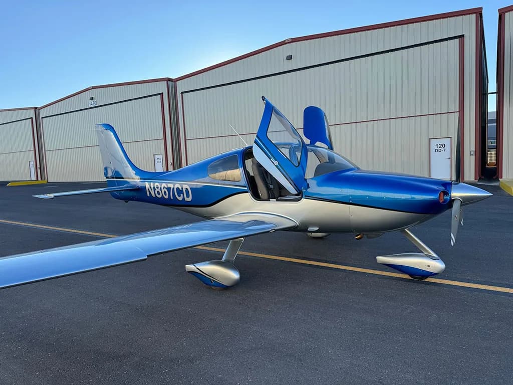 A blue and silver Cirrus SR22 aircraft with registration N867CD parked on an airfield with its doors open.