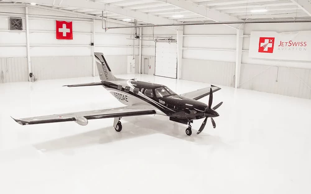 An aerial view of the black and silver Piper M600/SLS aircraft (N600AE) parked centrally in a spacious, white hangar.