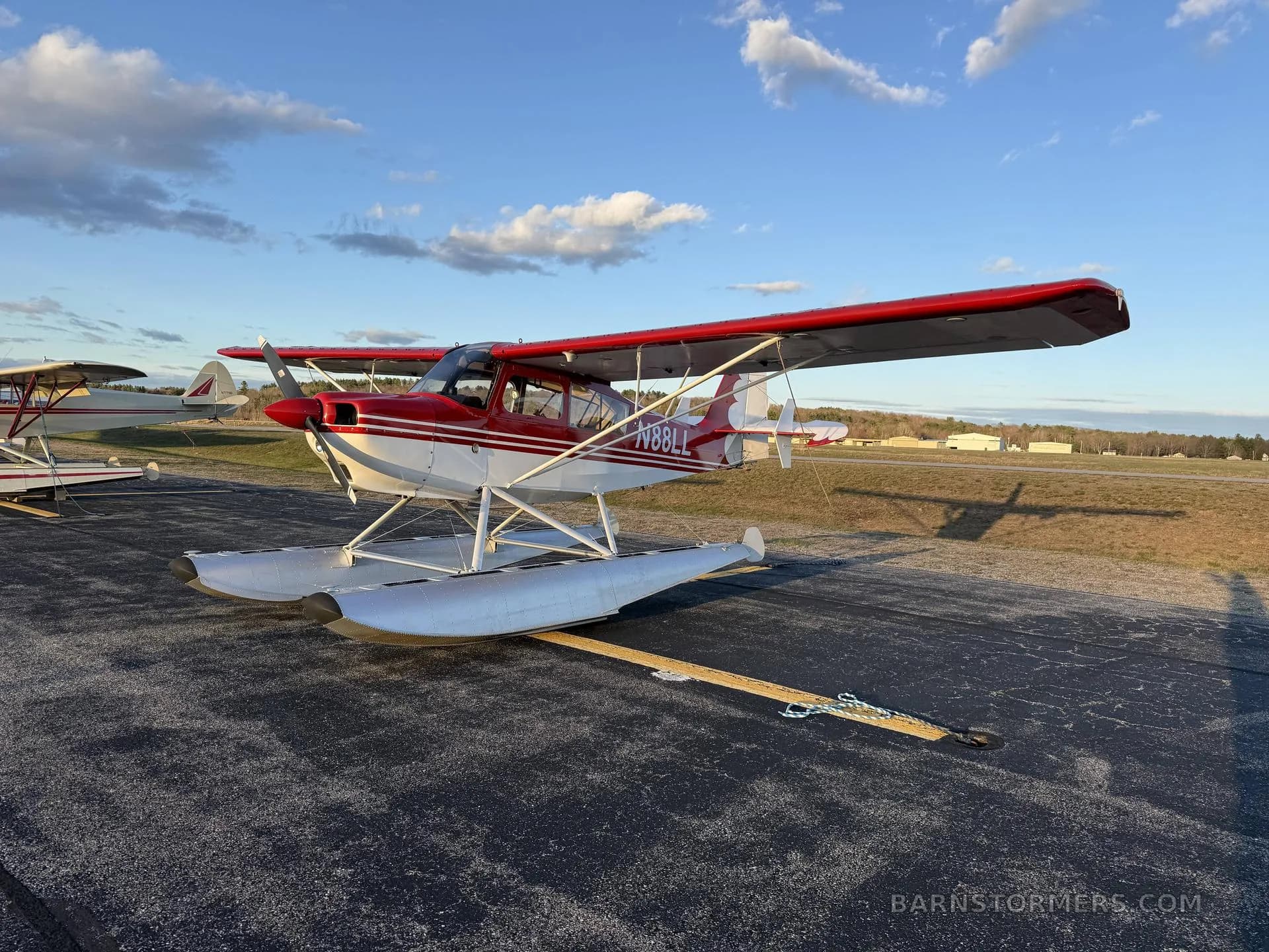 A red and white floatplane with tail number N88LL, parked on an asphalt runway with clouds in the sky.