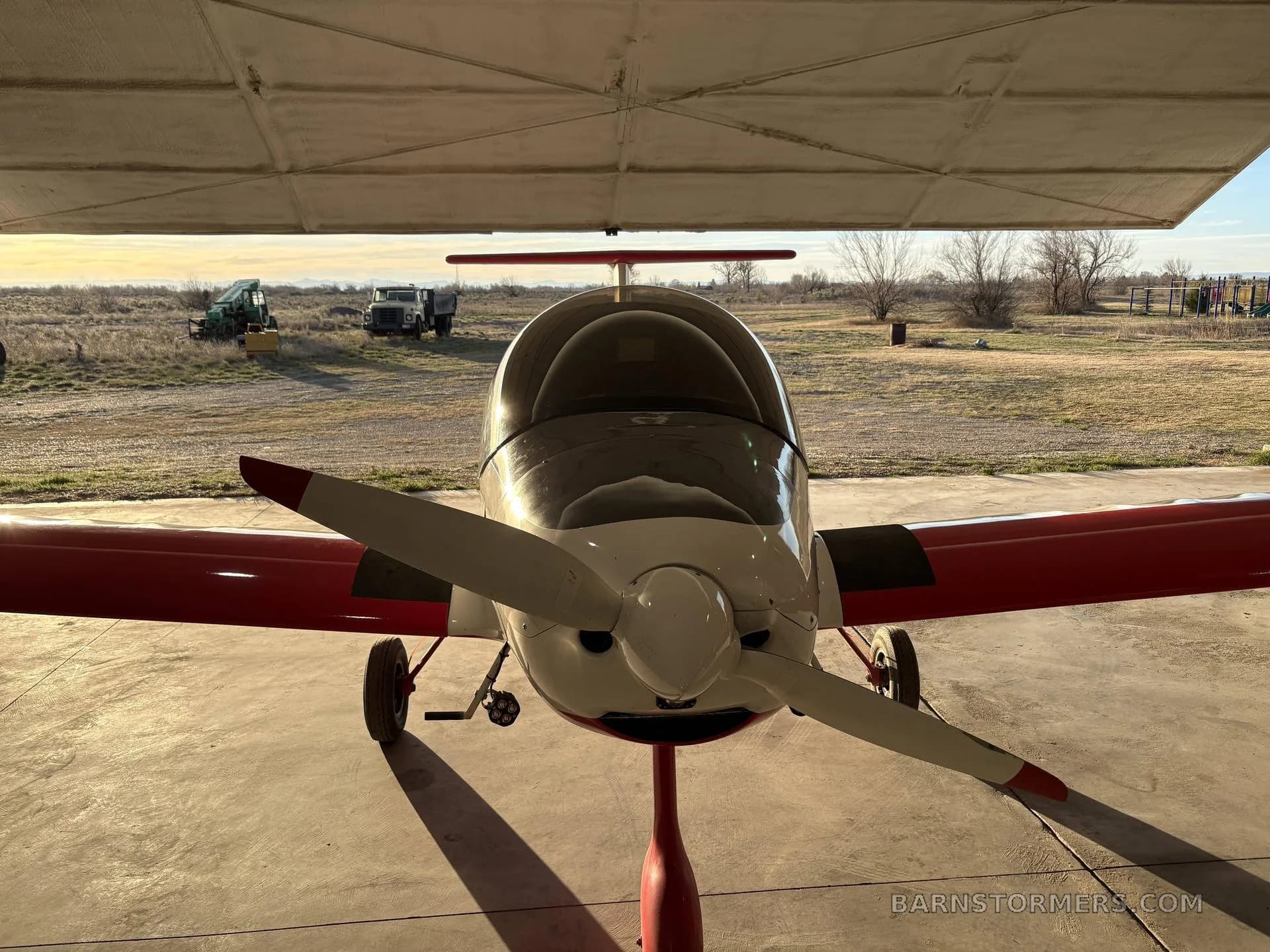 A white and red experimental aircraft, possibly an ATEC Zephyr, is parked head-on in a hangar with its propeller visible.