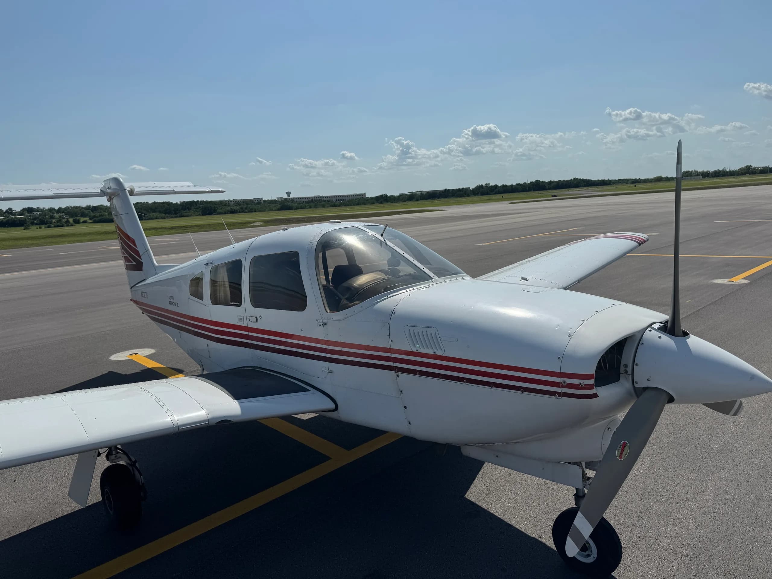A white Piper Arrow II aircraft with red and brown stripes parked on an asphalt tarmac, viewed from the front right.