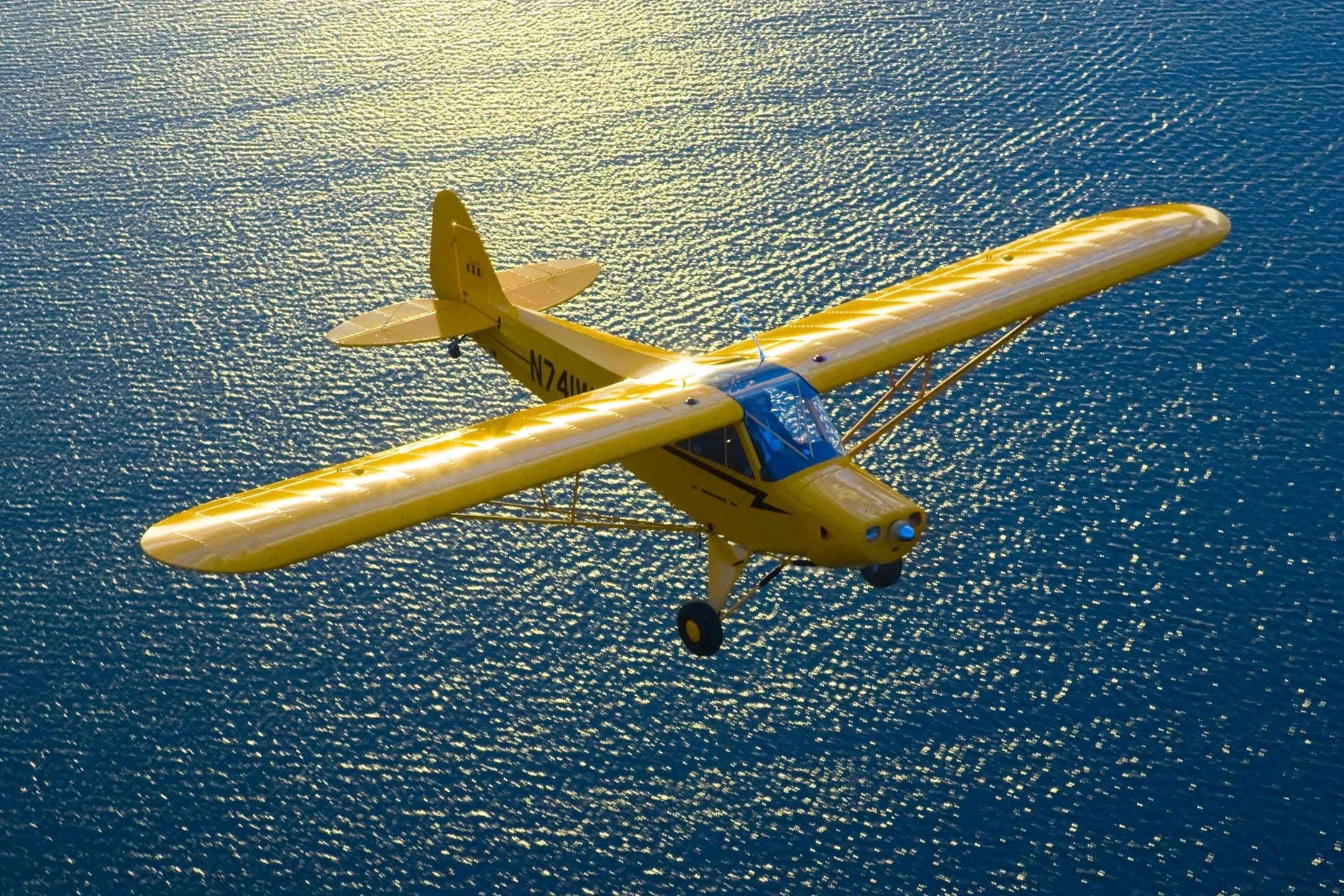 A yellow Piper Cub-style aircraft (N741W) flies over shimmering water with the sun reflecting brightly.