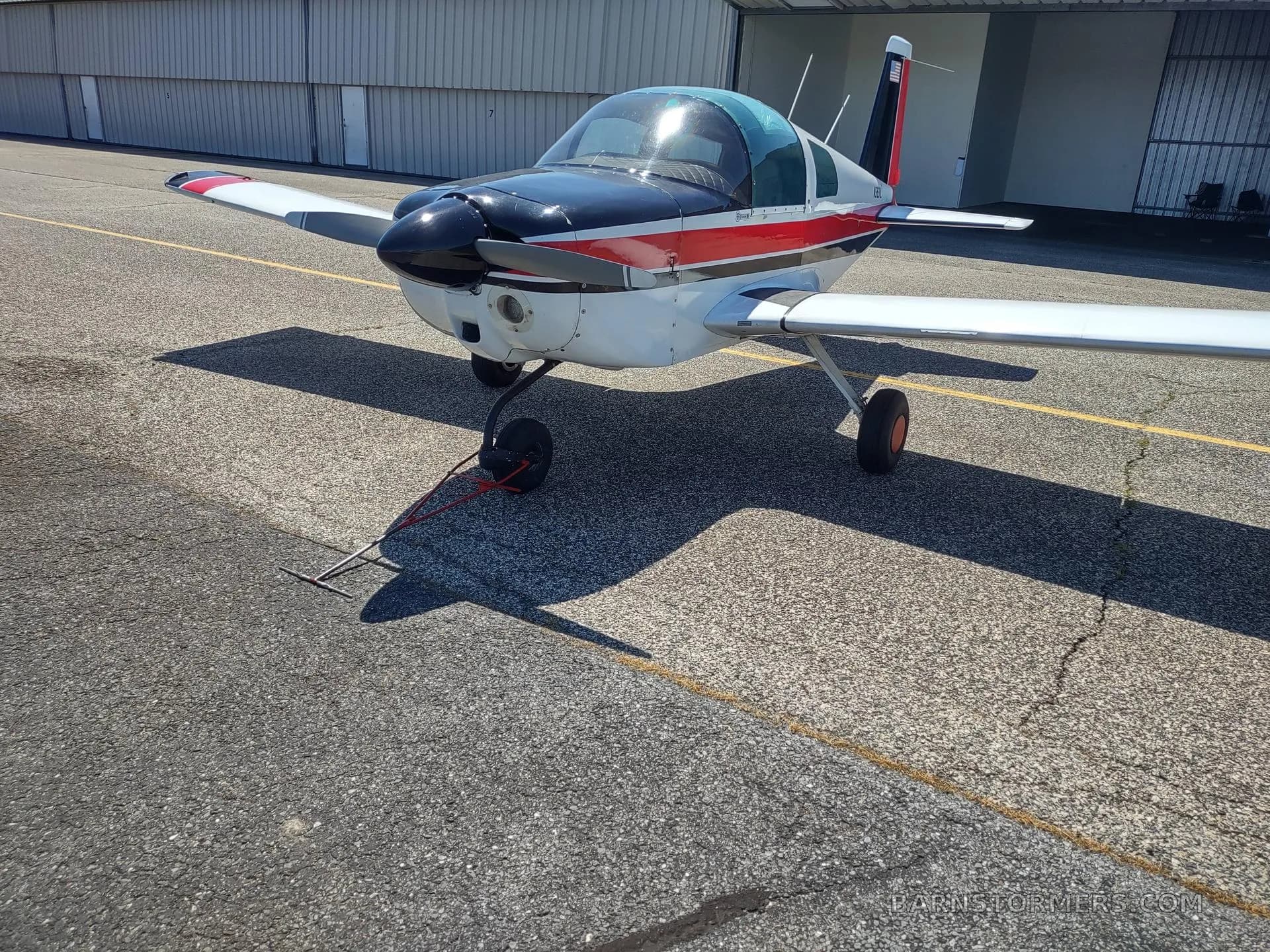 A front-left view of a white, red, and black single-engine aircraft with a bubble canopy on an asphalt ramp.
