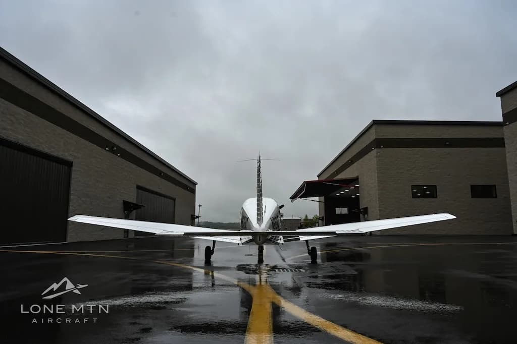 A white single-engine aircraft, N616AC, is viewed from the rear on a wet tarmac between two hangars.