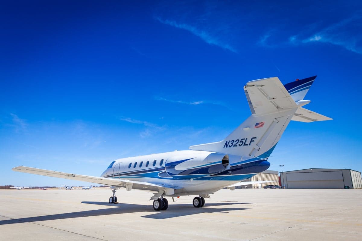 A white and blue jet sits on the tarmac in front of a hangar on a sunny day.