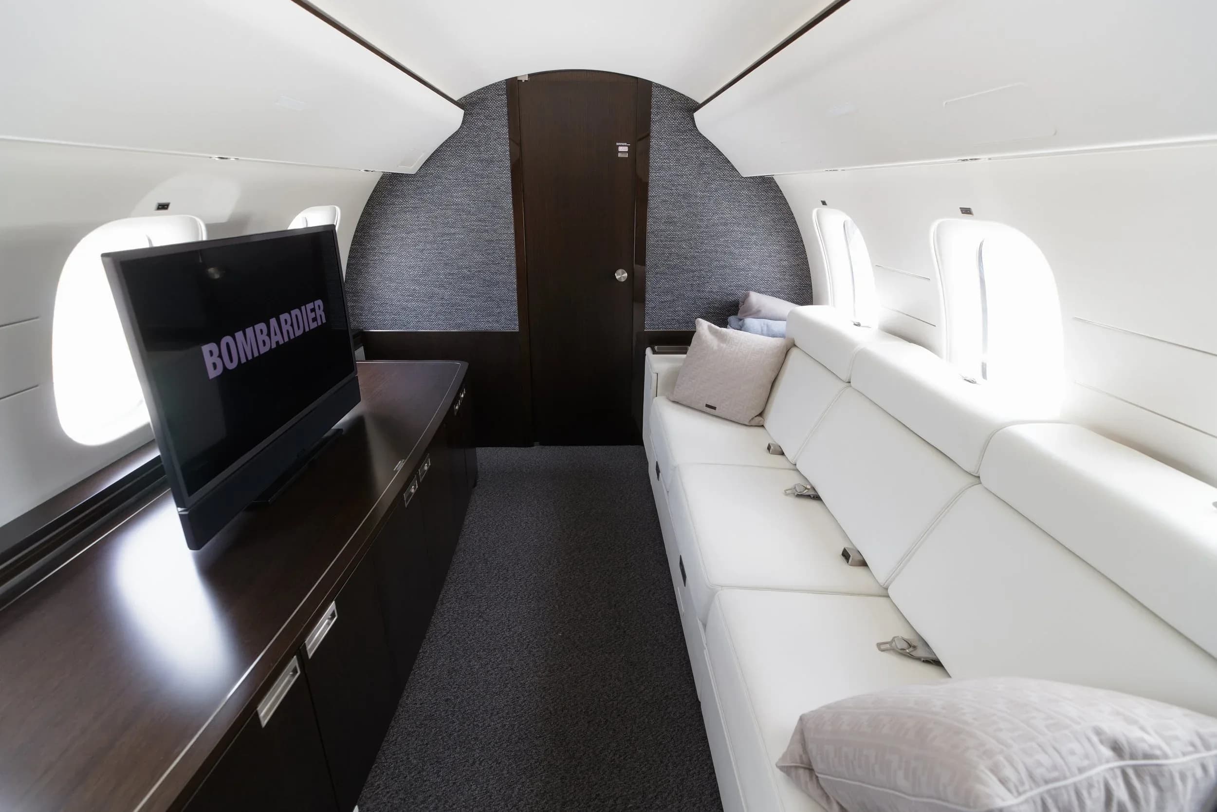 The aft section of a private jet cabin with a large screen displaying 'BOMBARDIER' and a comfortable white sofa.