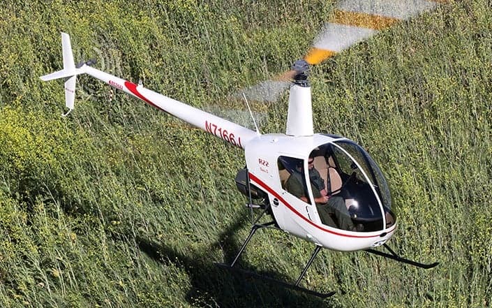 A white Robinson R22 helicopter flying over a grassy field.