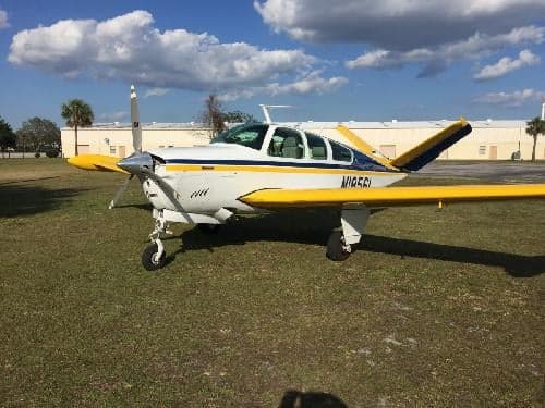 A Beechcraft Bonanza aircraft with a white, yellow, and blue paint scheme is parked on the grass.