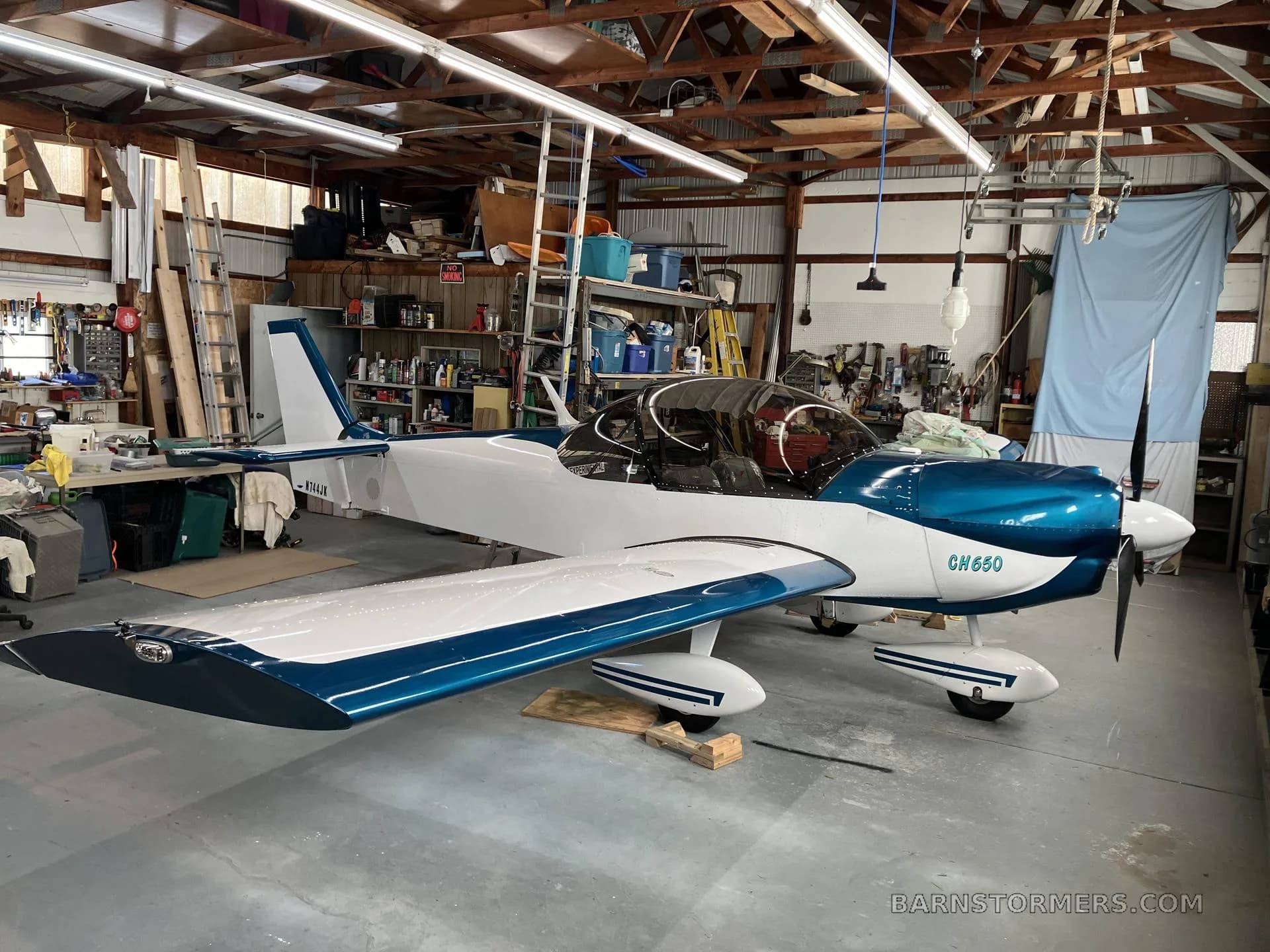 A white and blue Zenith CH650 aircraft with registration N744JK is parked inside a cluttered workshop.