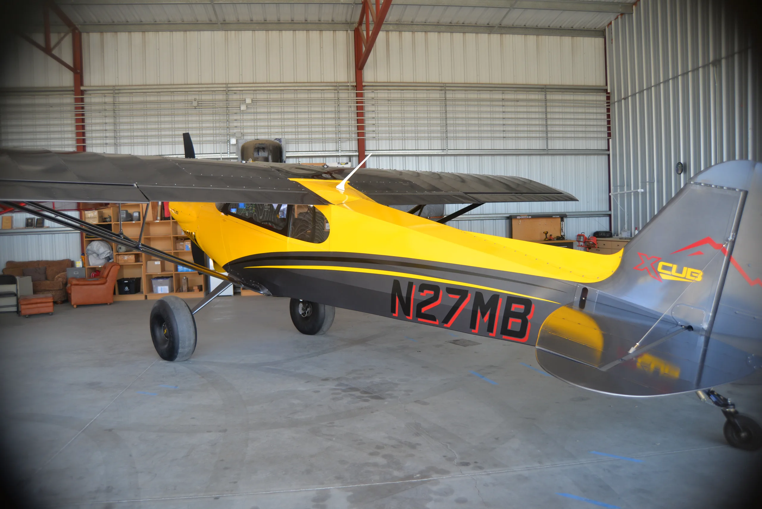 A yellow and gray CubCrafters XCub, registration N27MB, parked inside a hangar with various items in the background.