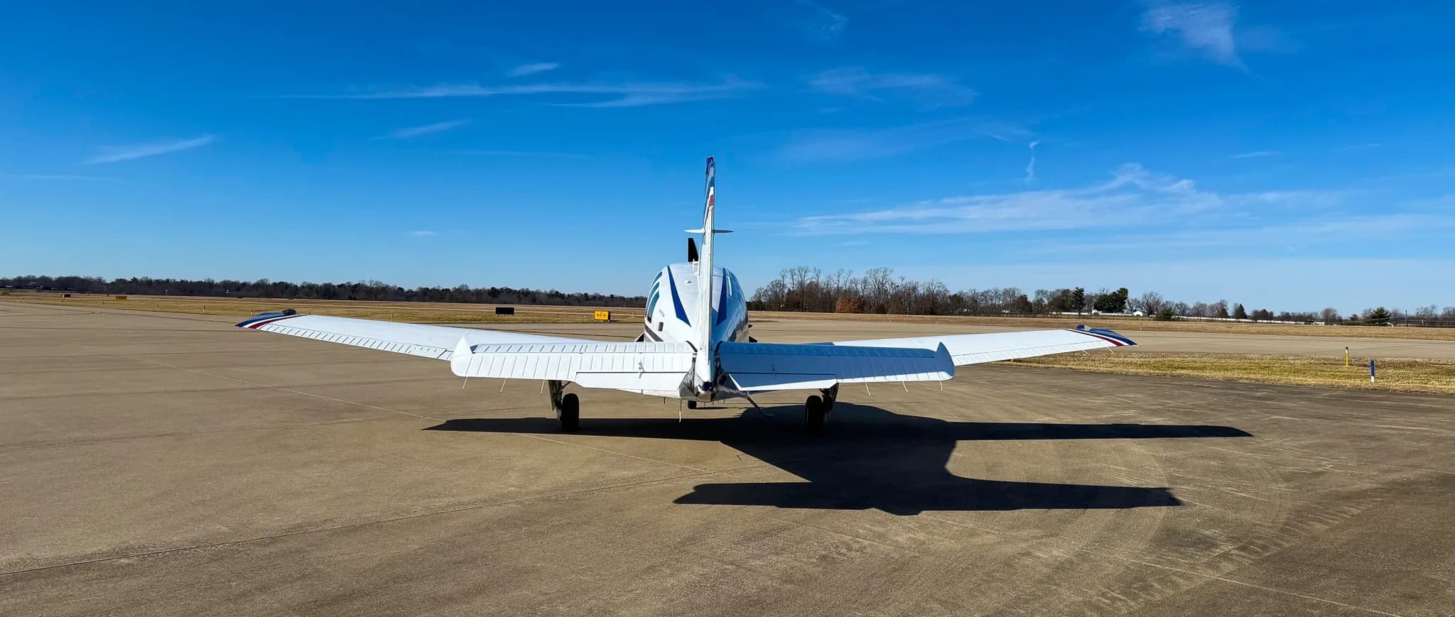 A rear view of a white Beechcraft Bonanza G36 with blue and grey stripes, registration N7CM, parked on an airfield.
