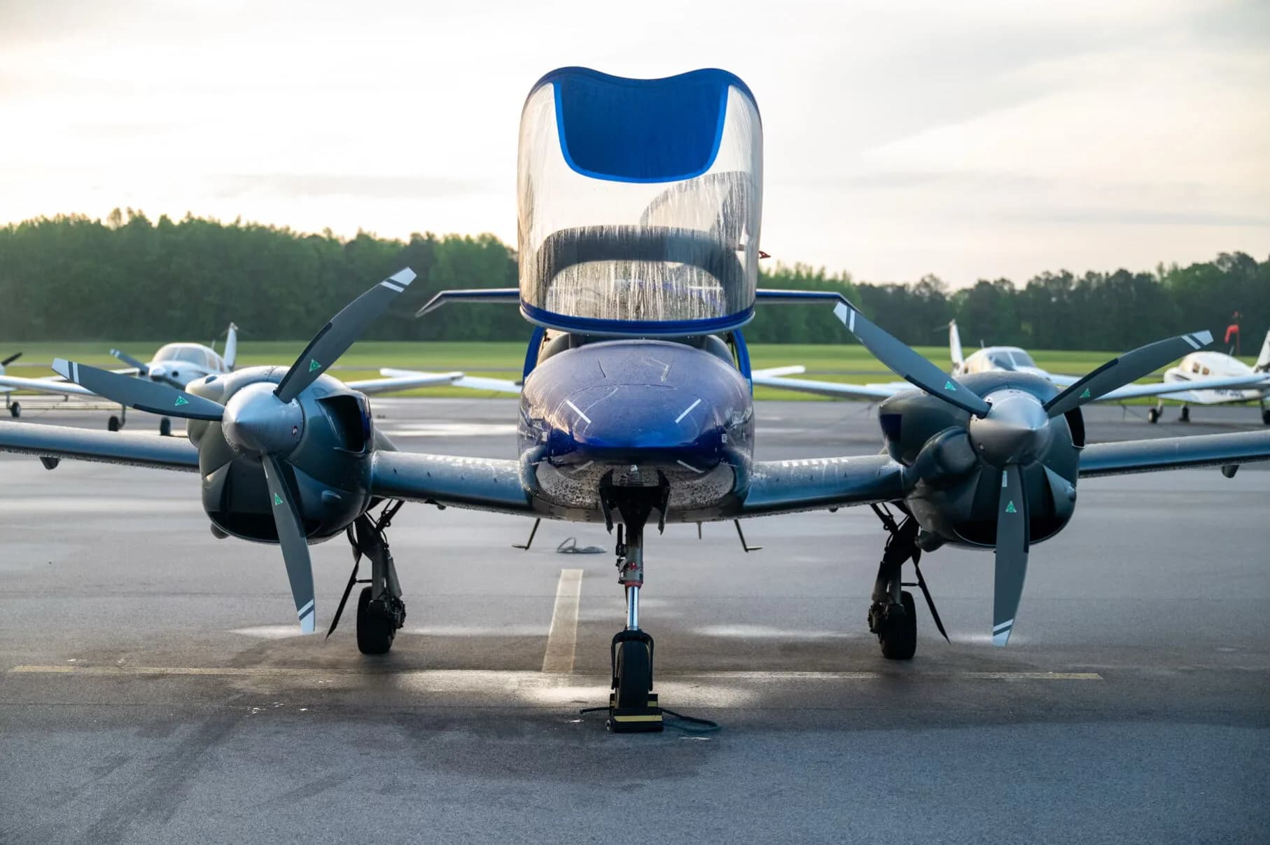 Close-up front view of the blue and grey Diamond DA42NG aircraft with its canopy open, showing water droplets on the surface.