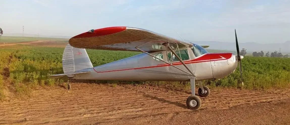 A silver and red Cessna 170 aircraft is parked on a dirt field.