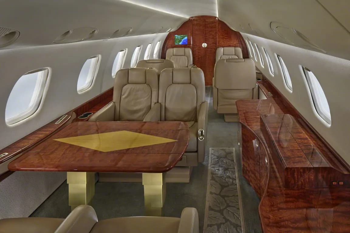 The interior of a private jet cabin featuring beige leather seats, a wooden table, and wood paneling.