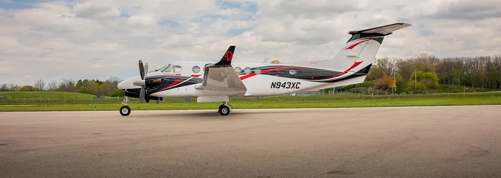 A white Beechcraft King Air 350 with black and red accents and tail number N943XC is parked on an airfield, viewed from the side.