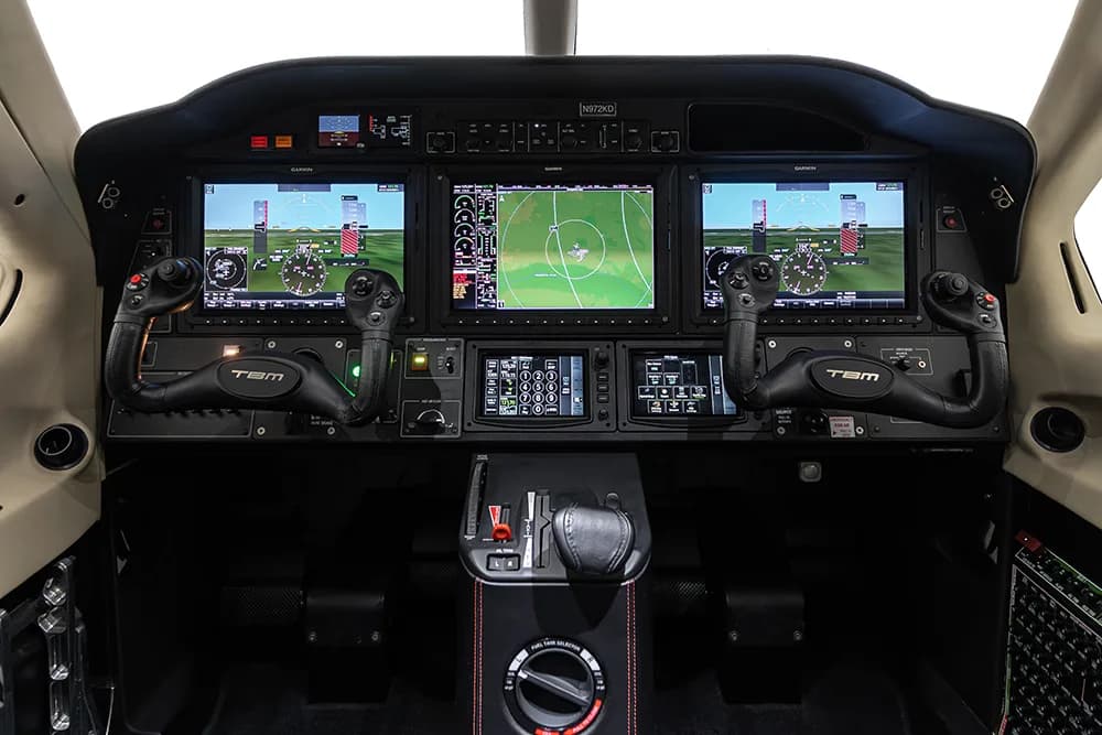 The cockpit of a TBM aircraft featuring a modern glass panel with three large displays and two yokes.