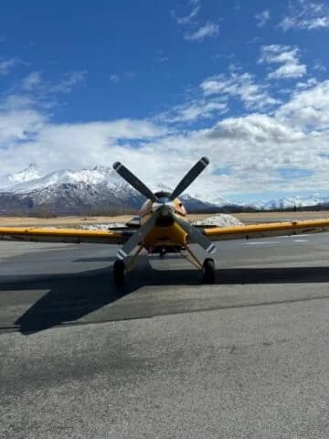 Front view of a yellow crop duster aircraft with a four-bladed propeller on an airfield, with snowy mountains in the background.