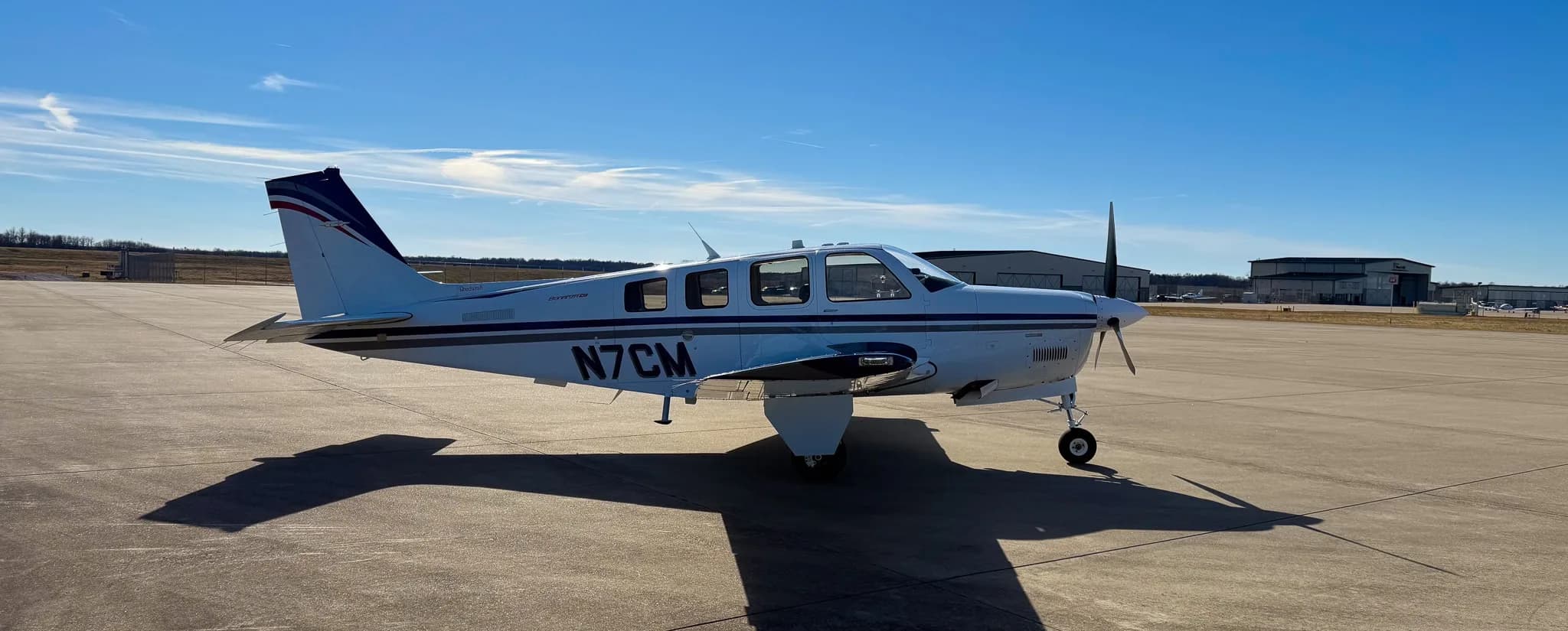 A white Beechcraft Bonanza G36 with blue and grey stripes, registration N7CM, parked on an airfield with hangars in the background.