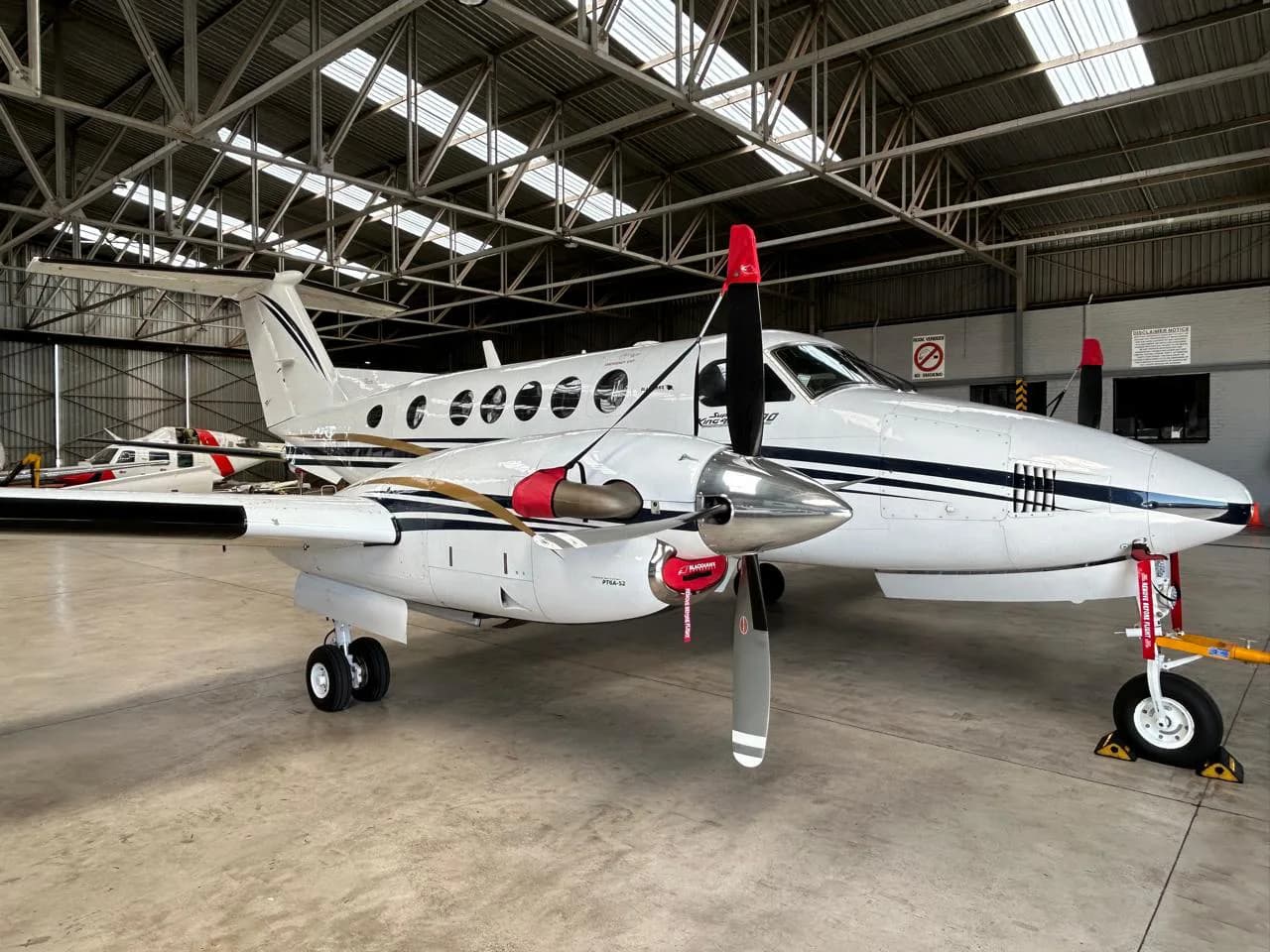 A white and black Beechcraft King Air aircraft is parked inside a hangar, with red propeller and pitot tube covers.