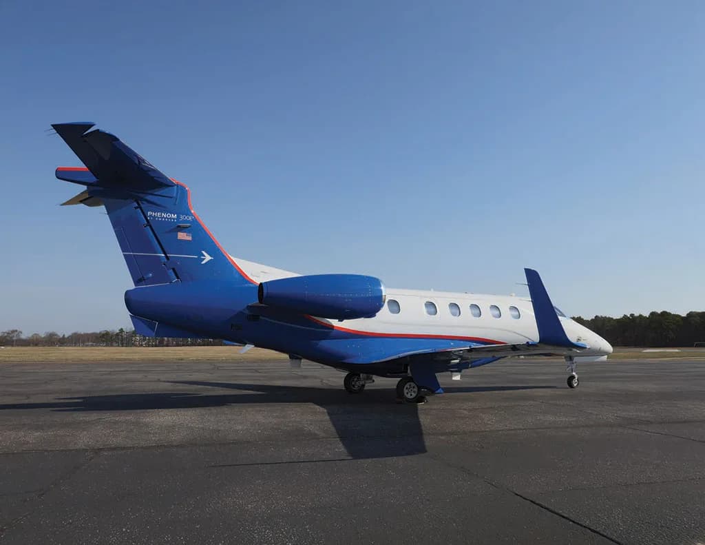 A white and blue Embraer Phenom 300 business jet is parked on an asphalt runway, viewed from the rear-right.