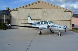 A Beechcraft Bonanza sits on the tarmac in front of a hangar.