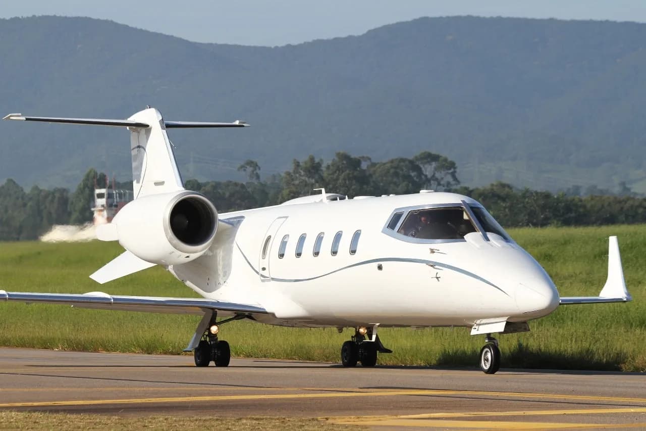 A white business jet with blue pinstripes is seen from a front-side angle on a runway with green grass and hills.