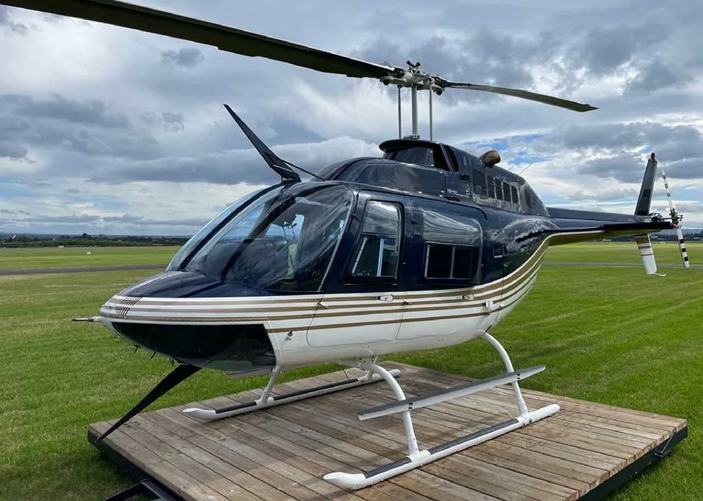 A black and white helicopter with gold stripes sits on a wooden platform on a grassy field under a cloudy sky.