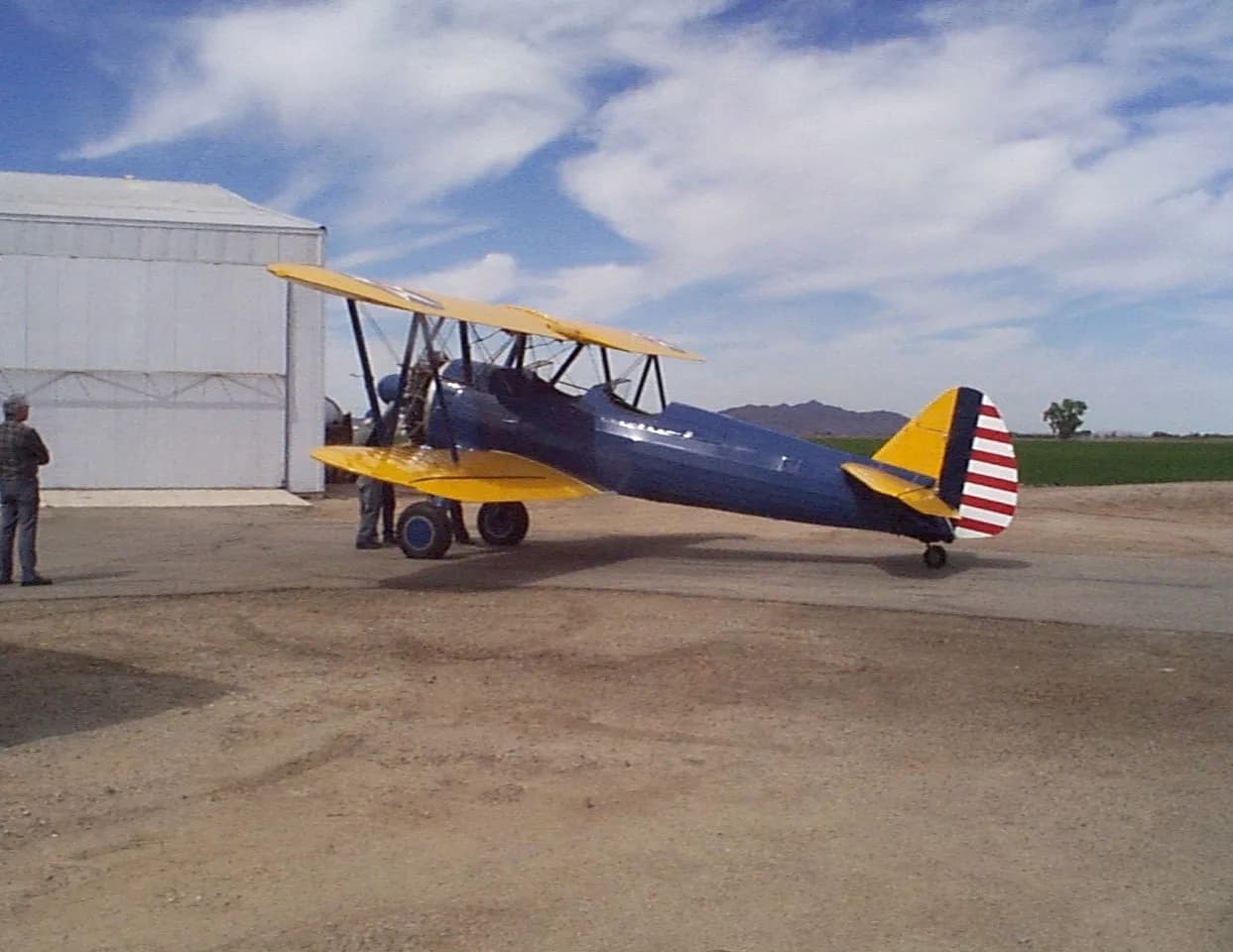 A blue and yellow biplane with a striped tail parked on a dirt and asphalt surface next to a white hangar.